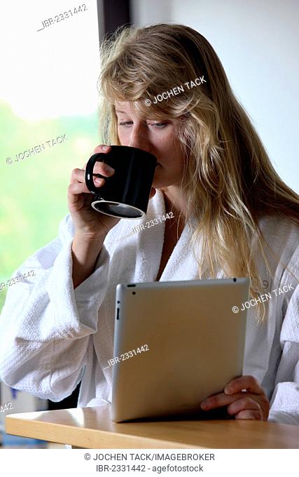 Young woman wearing a dressing gown drinking a cup of coffee in the morning, sitting in the kitchen reading an online newspaper on an iPad, a tablet computer