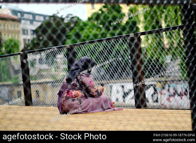 15 June 2021, Berlin: A woman in a headscarf looks at a smartphone while sitting on a wooden terrace on Maybachufer. Photo: Stefan Jaitner/dpa