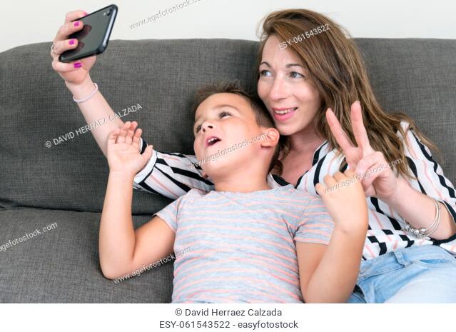 Cheerful mother holding smart phone making selfie with her little son showing victory sign. mom and kid boy sit on couch using gadget having fun taking...