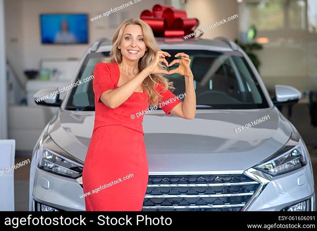 Happy moment. Young adult woman in red dress showing heart sign standing near new car smiling at camera in dealership