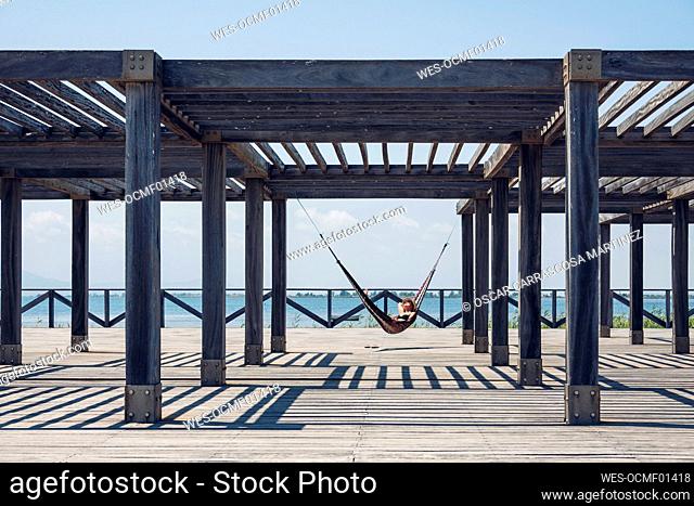 Woman lying in hammock hanging from metallic structure on boardwalk during sunny day