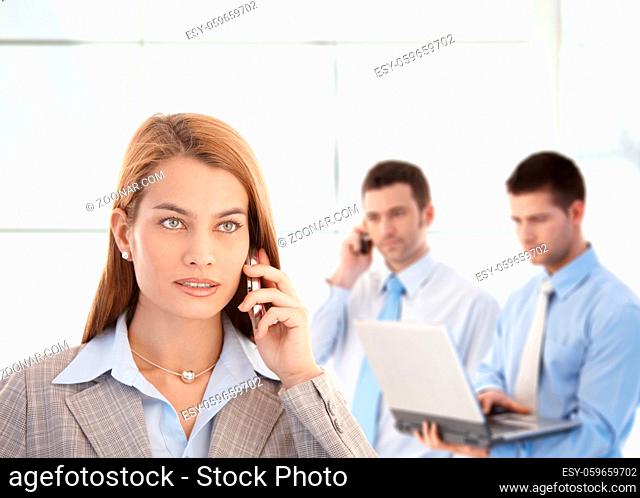 Attractive businesswoman talking on mobile phone, colleagues working in background
