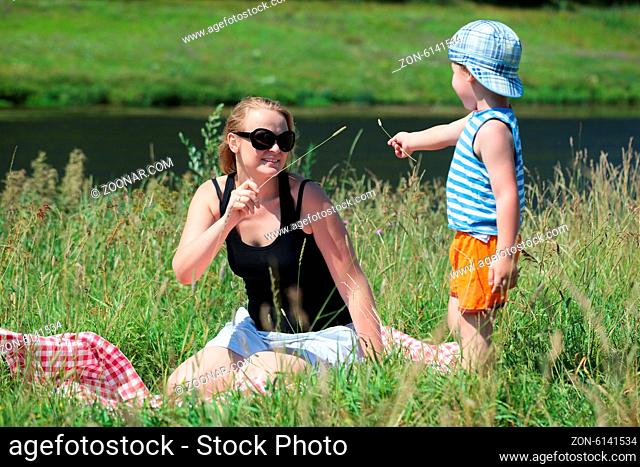 Happy mother and little son playing with grass on the meadow on the bank of a small river. Family fun outdoor