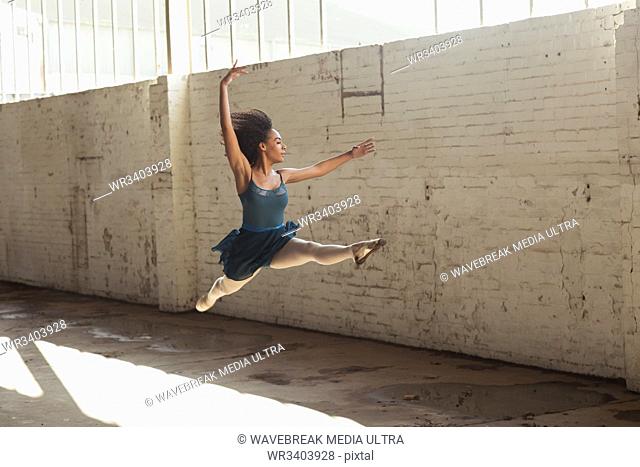 Side view of a young mixed race female ballet dancer leaping in the air with arms raised while dancing in an empty room at an abandoned warehouse