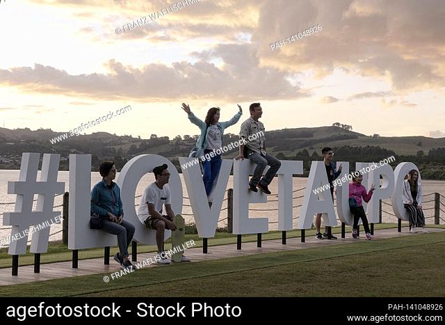 Big letters made of wood or plastic on the shores of Lake Taupo (crater lake, volcanic lake, caldera) that form the term #LOVETAUPA; sitting on it and waving...
