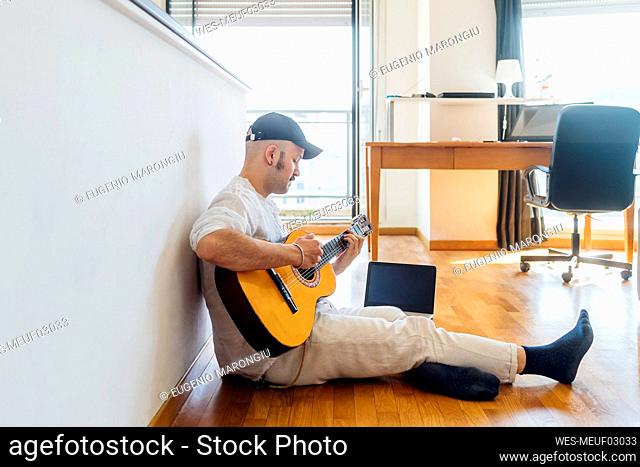 Male guitarist playing guitar by laptop while sitting on floor at home