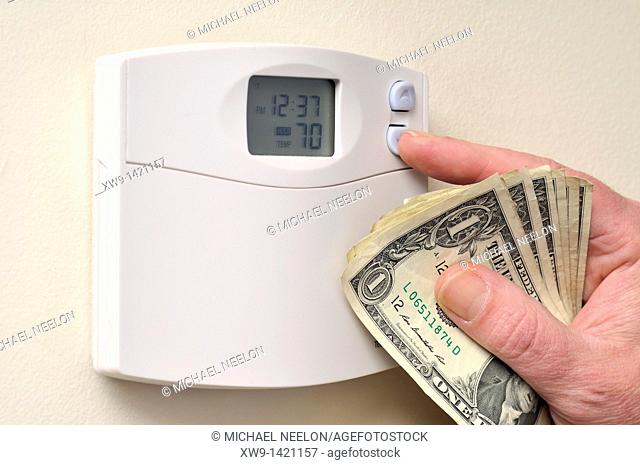 A hand clutching a some US dollar bills as it turns down the temperature setting heat thermostat