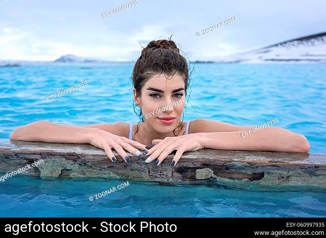 Pretty girl in a white swimsuit holds her hands on a wooden crossbeam in the geothermal pool on the background of snow mountains and cloudy sky outdoors in...