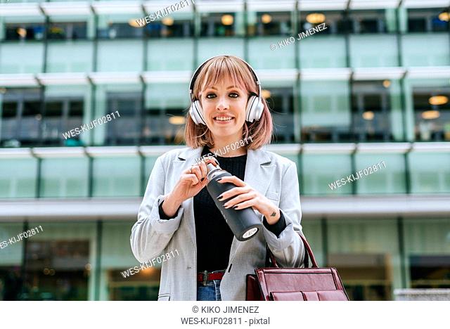 Portrait of smiling woman with headphones opening reusable bottle in the city