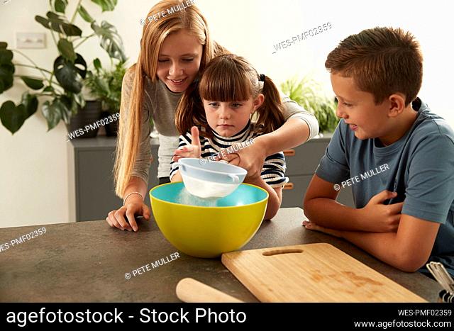 Smiling girl helping sister in sieving flour by brother at home