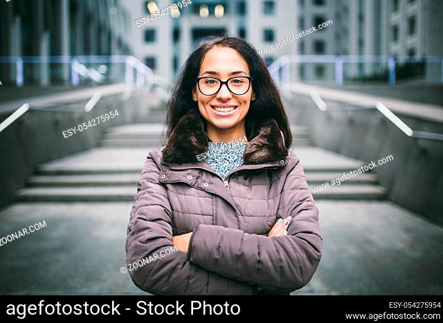 Beautiful young woman of European ethnicity with long brunette hair with a toothy smile, wearing glasses and a coat stands against backdrop of a business center...