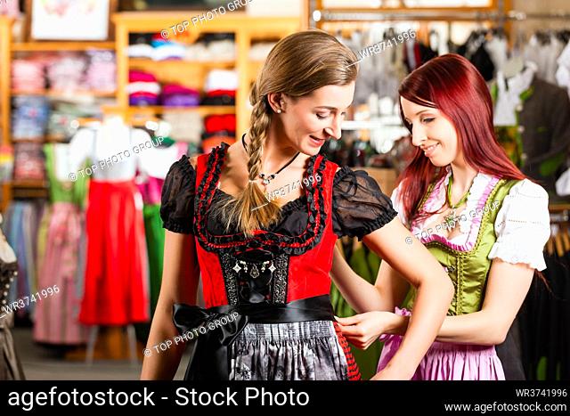 Traditional clothes - young woman is buying Tracht or dirndl in a shop, she has to try it on before and a friend is tying the ribbon