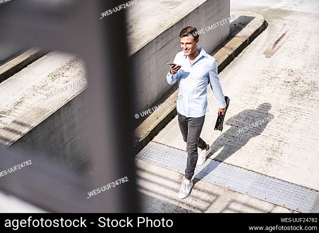 Young businessman with briefcase talking on smart phone through speaker during sunny day