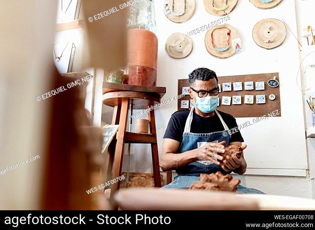 Potter wearing mask holding clay while sitting in workshop