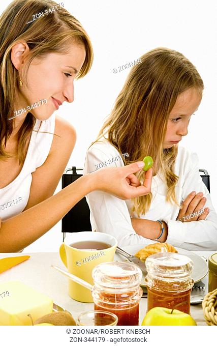 Mädchen sitzt mit verschränkten Armen von ihrer Mutter abgewendet am Frühstückstisch, weil diese ihr Weintrauben anbietet