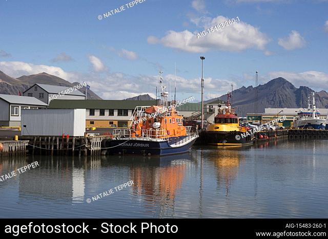 A busy fishing harbour in Hofn, Iceland, with boats moored outside the dock and mountains in the background