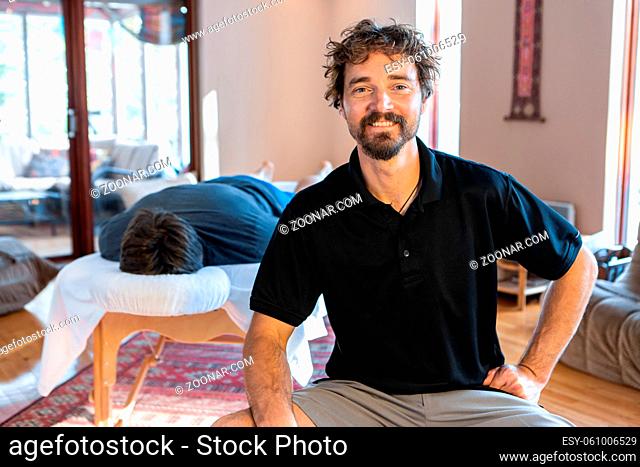 Portrait of smiling physiotherapist on working place. A strong male client lying on the table and waiting for treating body massage