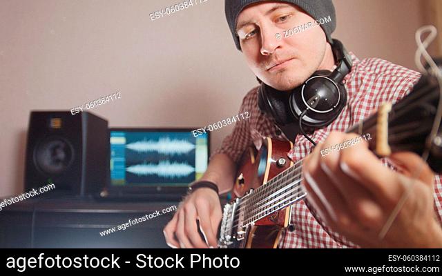 Young male musician composes and records soundtrack playing the guitar, using computer, headphones and keyboard, focus on equipment