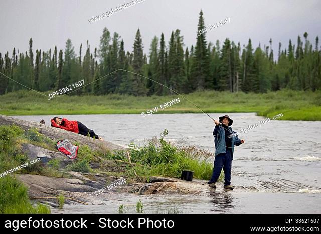 A woman lounges on a rock while her friend casts his fly rod.; Winisk River, Ontario, Canada