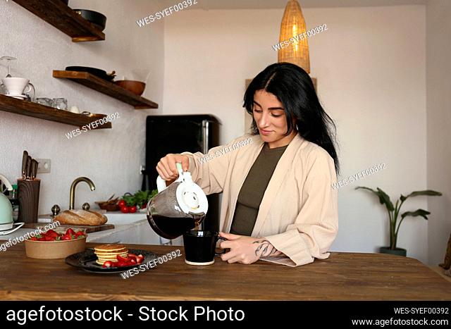 Young woman pouring freshly brewed coffee in cup at kitchen table