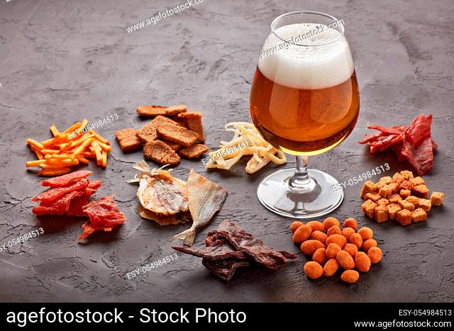 Glass with beer and set of various kind snacks: jerky, potato chips, salted nuts, stockfish, bread spicy crackers, dried squids on dark gray background
