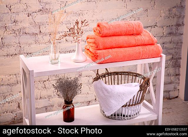 Rack with a stack of three peach color towels and baskets with clean white towels and toilet decor near a brick wall. Shelf with terry towels and decorative...