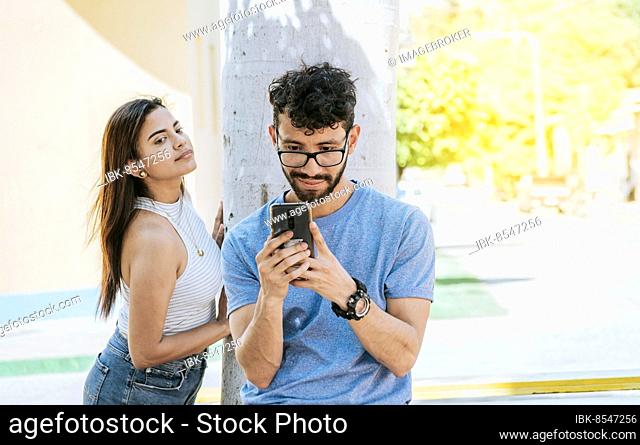 Jealous girl spying on her boyfriend's cell phone in the park, Distrustful woman spying on her boyfriend's cell phone outdoors