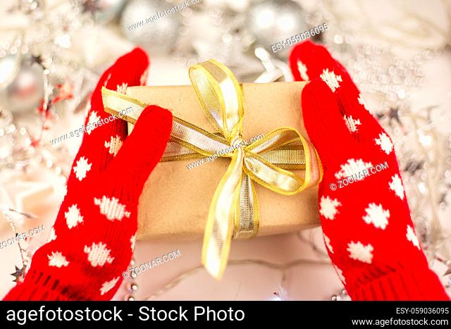 Female hands hold a Christmas gift with a gold ribbon in red mittens, new year mood, preparation for the celebration