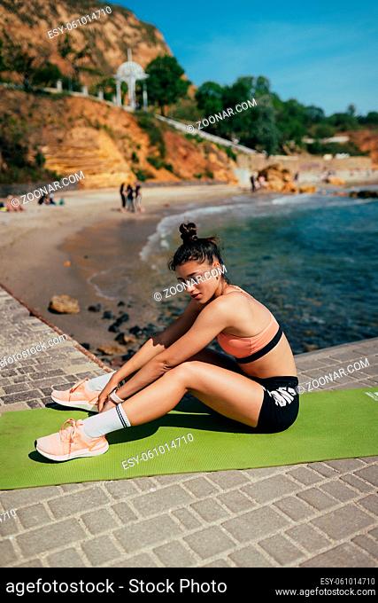 Young brunette woman wearing fitness outfit, stretching on yoga mat outside on pier in summer. Fit girl, doing yoga poses by sea