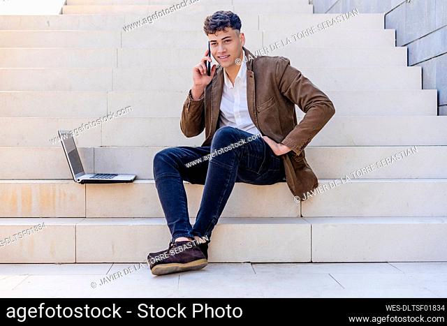 Male entrepreneur sitting with laptop while talking on mobile phone at staircase