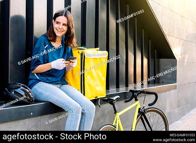 Smiling delivery woman with smart phone sitting on wall