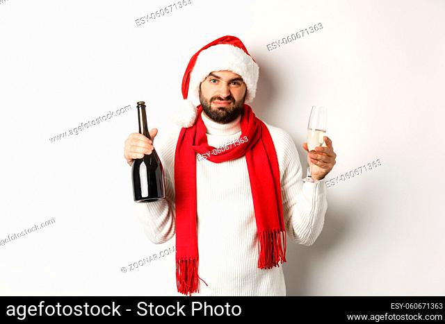 Christmas party and holidays concept. Skeptical bearded man in Santa hat and scarf, holding champagne and complaining, standing against white background