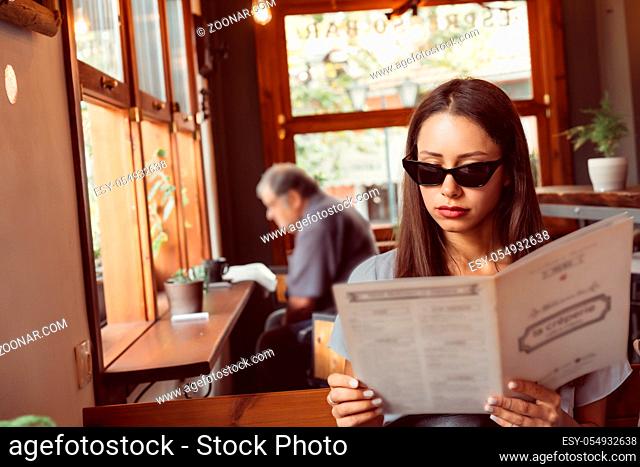 Attractive woman in sunglasses is sitting in a restaurant. The girl is reading the menu
