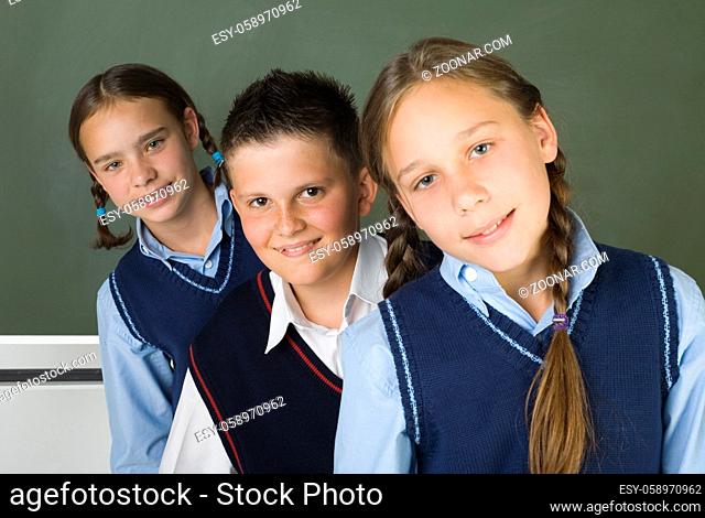 Portrait of three students: two girls and a boy in the middle of them. They're looking at camera and smiling. Blackboard behind them