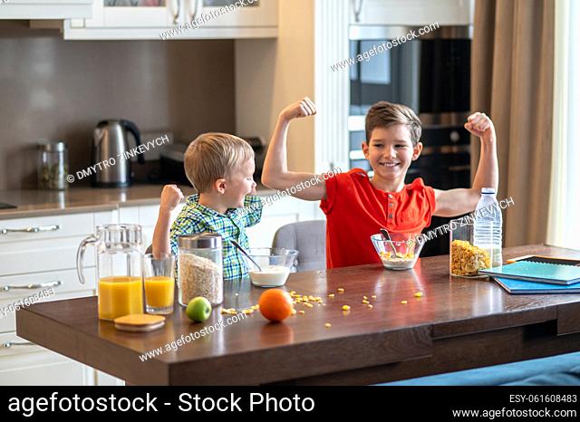 Smiling cute boy and his pleased younger brother flexing their muscles at the kitchen table