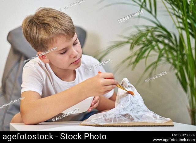 Boy with a brush in her hands glues a piece of paper for papier mache. Child makes a creative project in the form of a mountain for homework