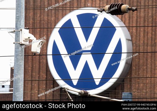 27 July 2020, Lower Saxony, Wolfsburg: A camera hangs at the Wolfsburg train station, while in the background the large VW logo can be seen on the facade of the...