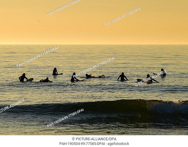 21 April 2019, Denmark, Klitmöller: Surfers are waiting for a wave in the water of the North Sea, while the approaching sunset bathes everything in a soft light