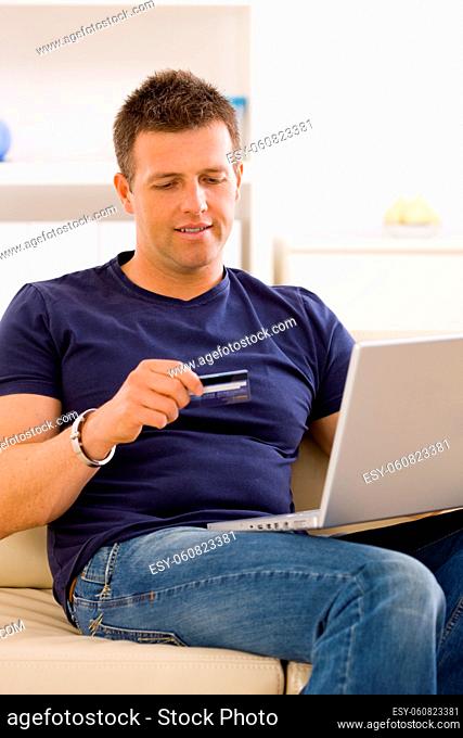 Young man shopping online at home using laptop computer, sitting on couch, looking at credit card