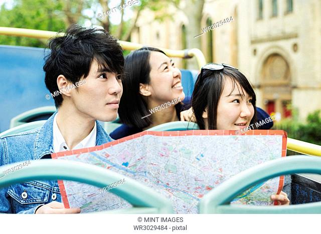 Smiling man and two women with black hair holding city map, sitting on the top of an open Double-Decker bus, driving along urban road