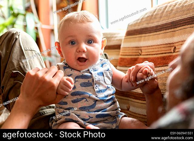 Portrait of cheerful and excited infant baby sitting on father lying on couch and holding hands while playing and jumping on stomach