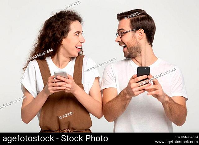Excited amazed young attractive couple looking at each other, holding cellphones in hands, received online win notice, playing internet games together