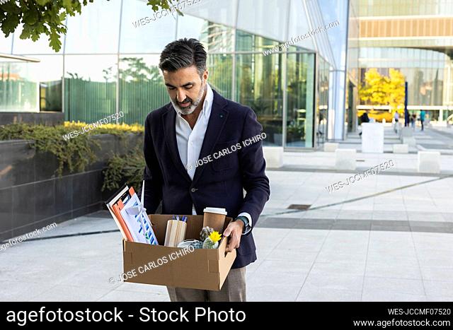 Sad businessman looking at office supplies in box standing near building