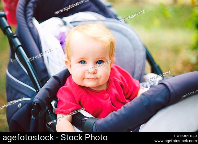 Portrait of a little funny child girl blond with blue eyes sitting in a baby stroller in the summer for greens. Trinasport for a child and transportation of...