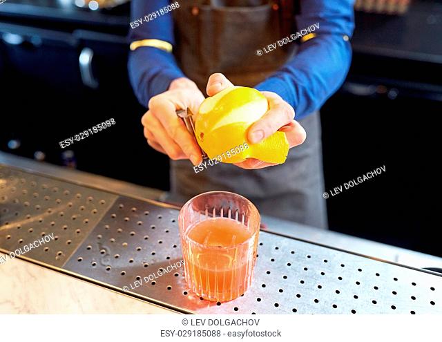 alcohol drinks, people and luxury concept - bartender with glass and peeler removing peel from lemon and preparing cocktail at bar