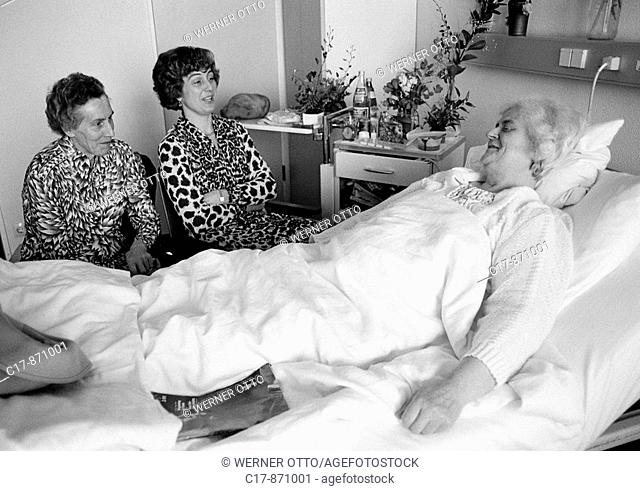Eighties, black and white photo, people, health, older woman lies in a sickbed of a hospital, two women visit her sitting beside the bed, aged 70 to 80 years
