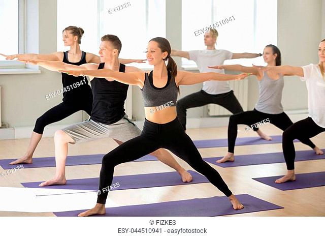 Group of young sporty people practicing yoga lesson, doing Warrior two exercise, Virabhadrasana 2 pose, , yogi students working out indoors in sport club