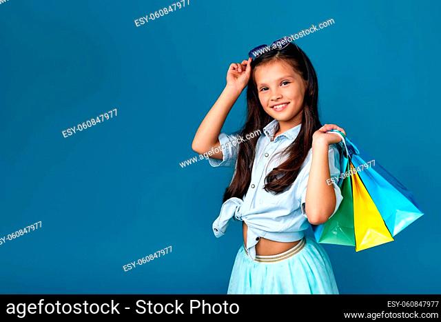 happy little caucasian child girl holds many shopping bags with purchases isolated on blue background. copy space