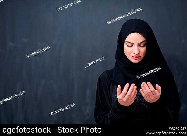 portrait of beautiful muslim woman in fashionable dress with hijab making traditional prayer to God keeps hands in praying gesture isolated on black chalkboard...