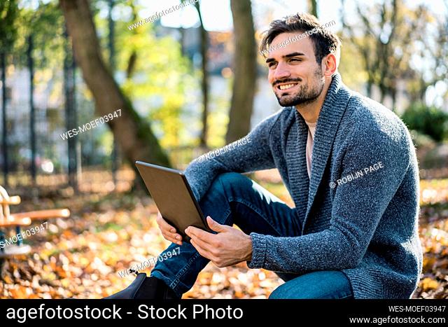 Thoughtful smiling young man holding digital tablet while sitting at park